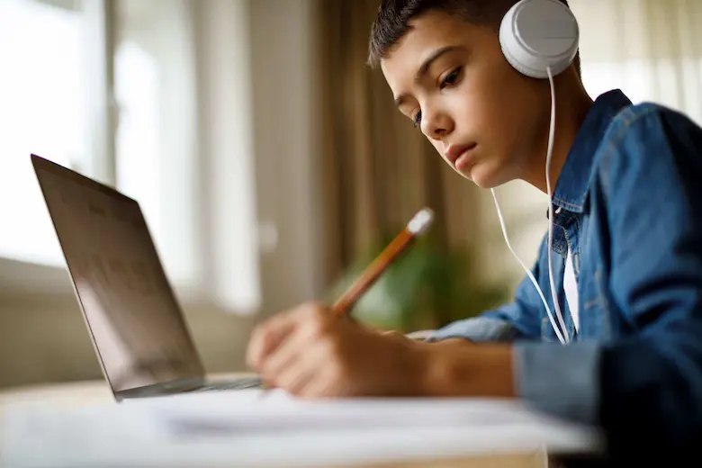 A focused boy wearing headphones while writing on a notebook, engrossed in online courses