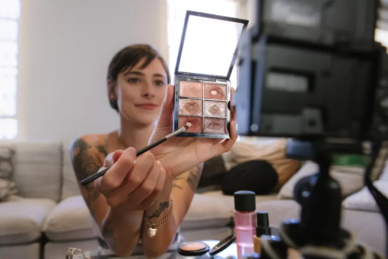 A women is holding up a makeup palette and brush, demonstrating in front of a camera. They are sitting on a couch with makeup products on a table in front of them