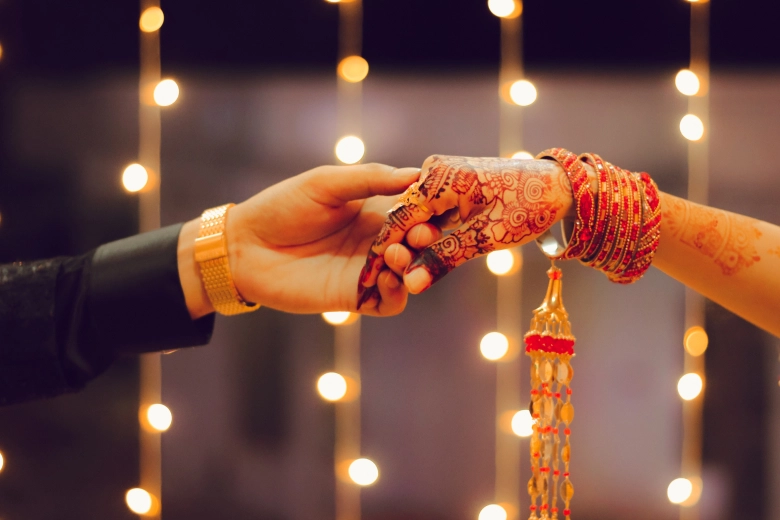 Close-up of a couple holding hands, adorned with henna and bangles, against a background of string lights.