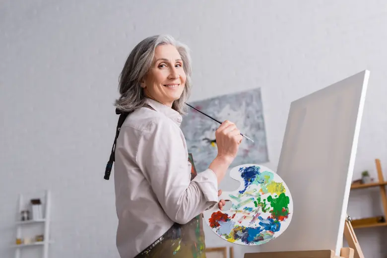 A smiling older woman with grey hair holds a paintbrush and a paint palette, standing in front of a blank canvas on an easel