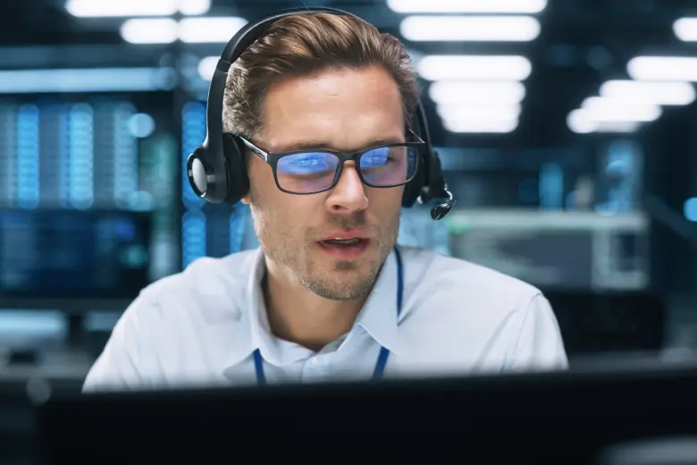 Man wearing glasses and a headset working on a computer in a modern office with multiple monitors in the background.