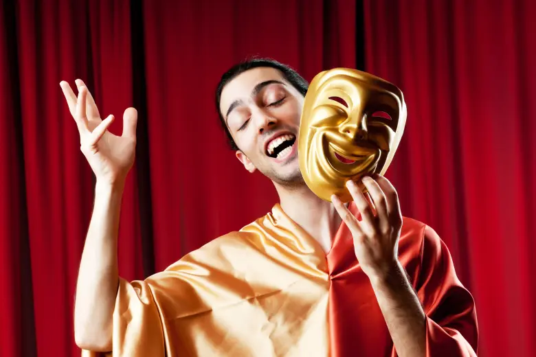 A person dressed in a colorful robe holding a golden comedy mask in one hand, smiling with their eyes closed, in front of a red curtain background.