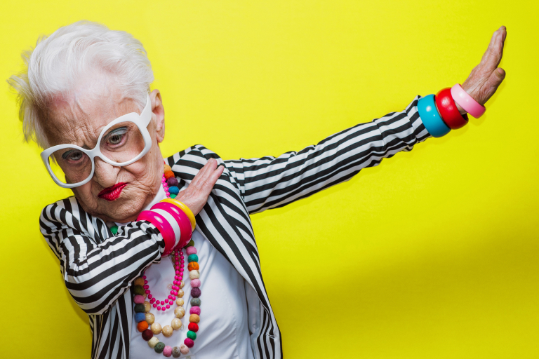 n elderly person with white hair, wearing white-rimmed glasses, a striped jacket, colorful bracelets, and a beaded necklace, poses against a yellow background with one arm dabbing.